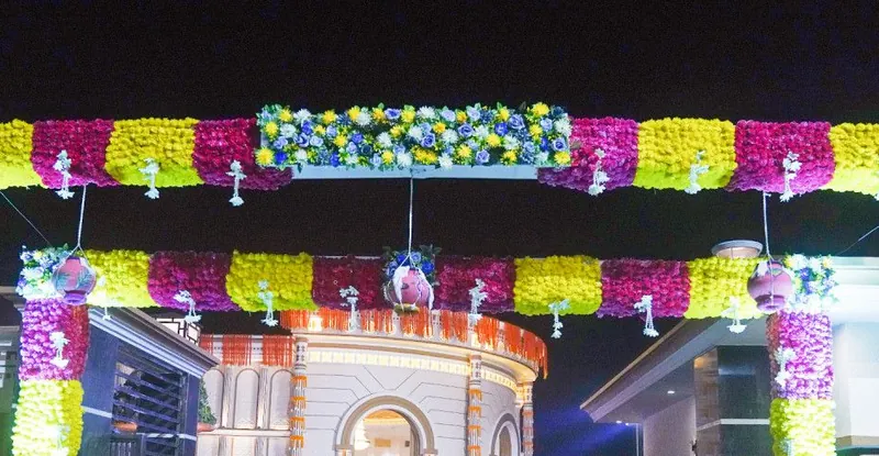 A gateway of white, yellow and pink flowers at night at Nurmahal Ashram, Janmashtami 2025, with a lit building behind it
