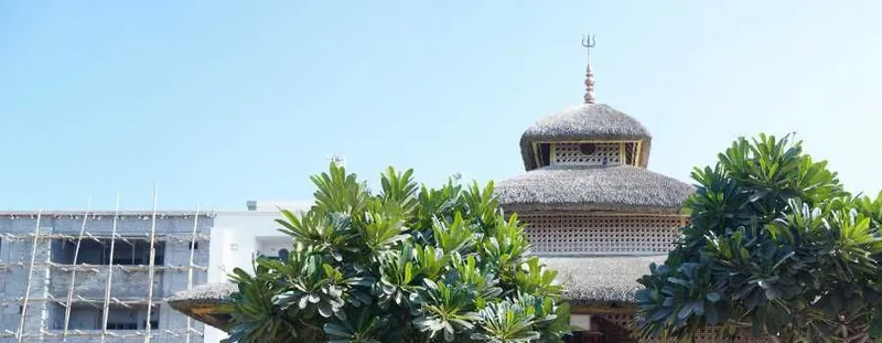 A tiered thatched pavilion between frangipani trees under a clear blue sky at Nurmahal Ashram